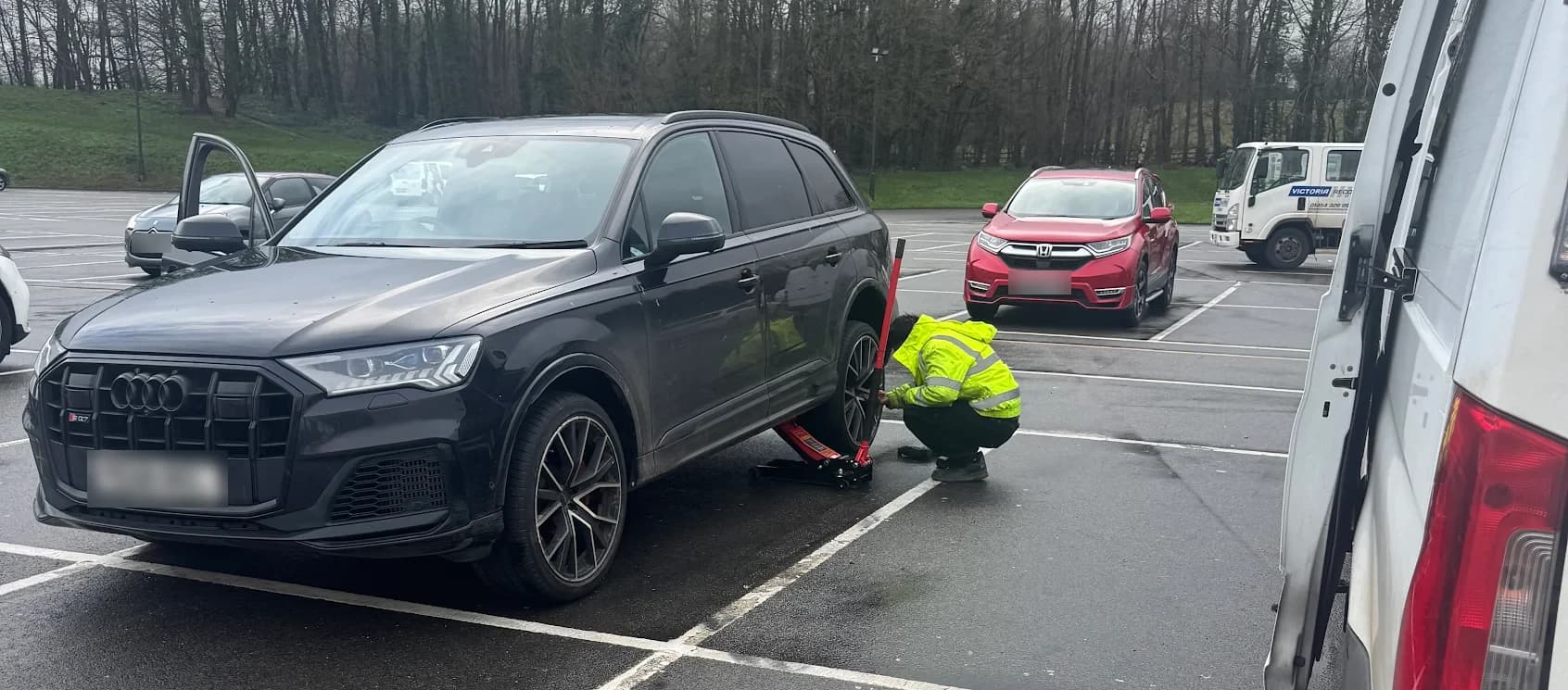 A fitter in a hi-vis jacket kneeling at the front wheel of a black Audi on a trolley jack in a car park, with the open door of the van in the foreground